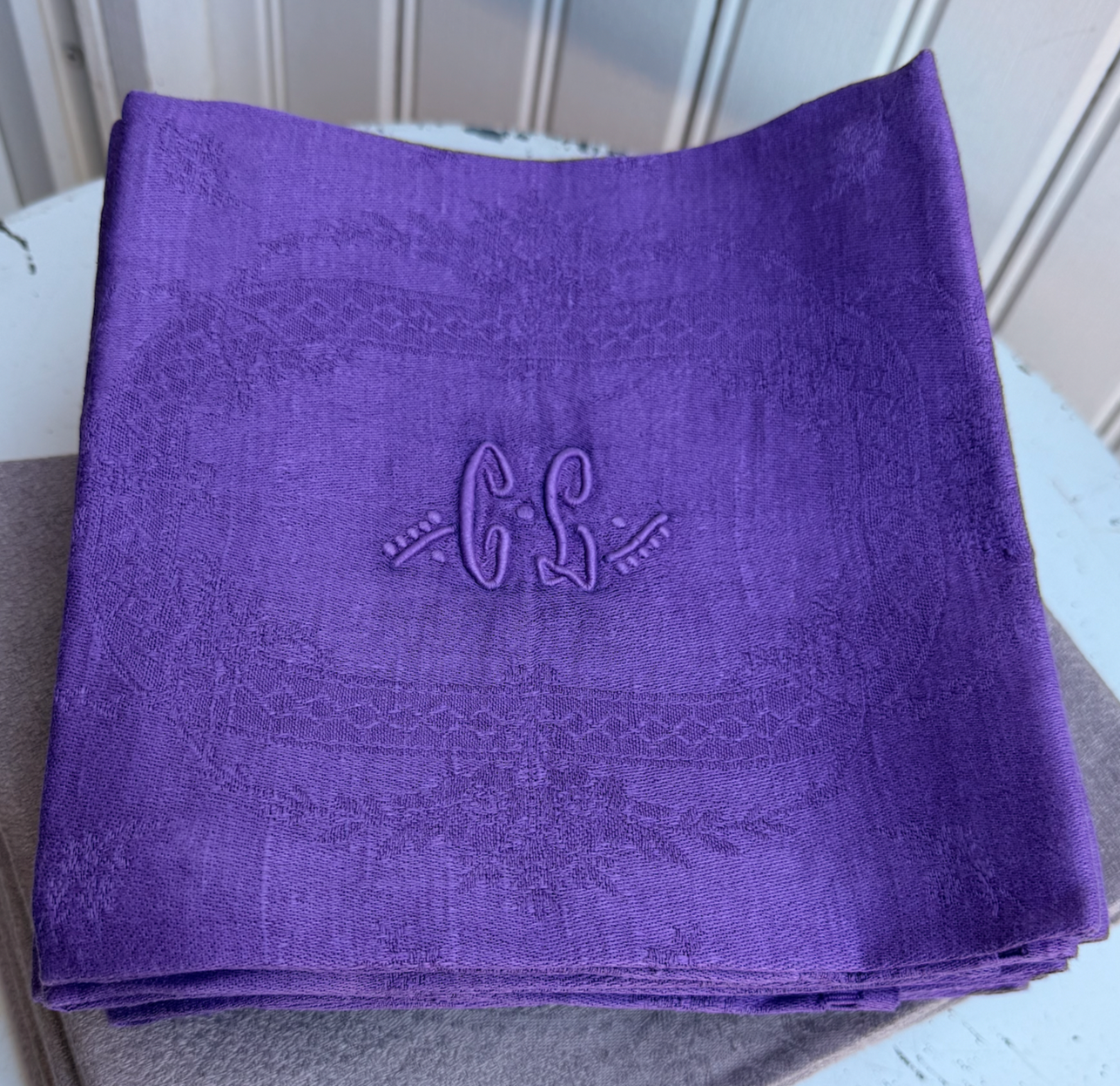A folded purple woven fabric napkin with a subtle damask pattern and an embroidered monogram 'C.L' in the center, displayed on a gray surface.